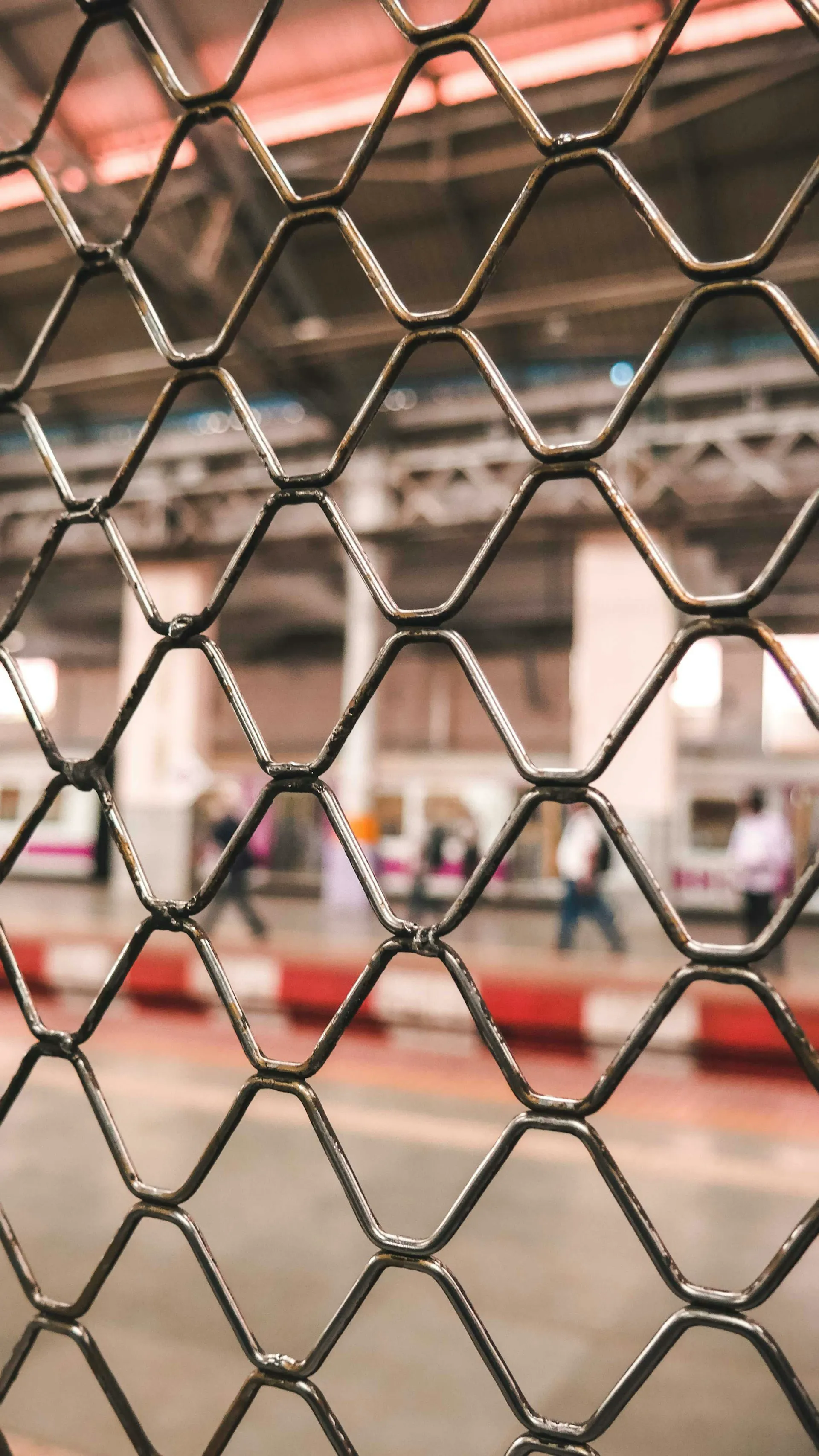 Een metalen hekwerk met een zeshoekig patroon, op de achtergrond zijn mensen en een trein perrong zichtbaar